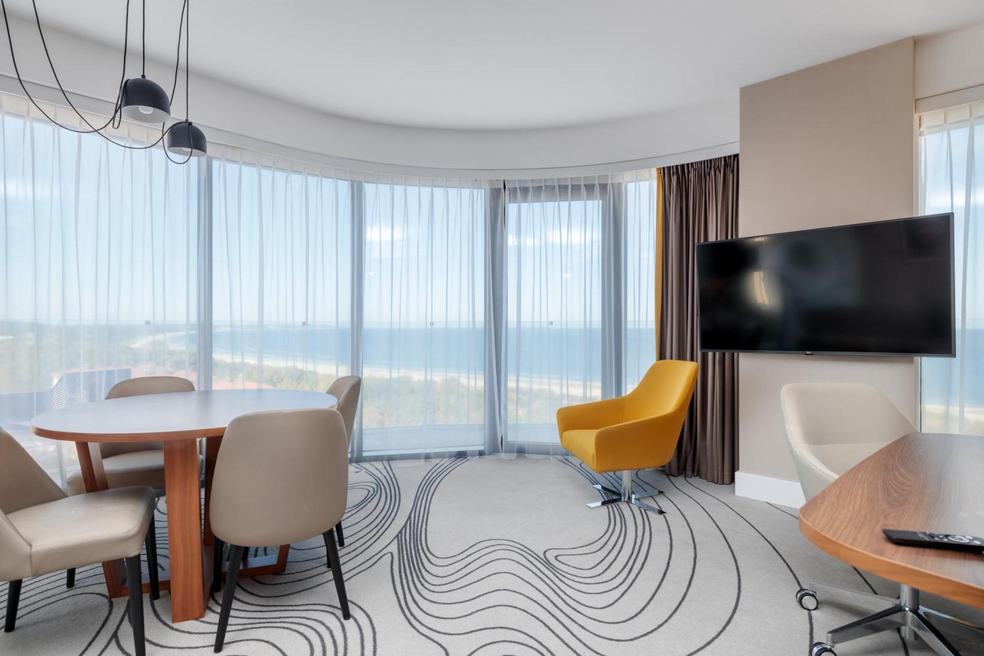Radisson Blu Resort 슈비노우이스쳬 외부 사진 The image shows a modern hotel room or suite with a bright, airy atmosphere. There are large windows that allow natural light to fill the space, providing a view of the ocean. The decor includes a round dining table with several chairs, and a stylish