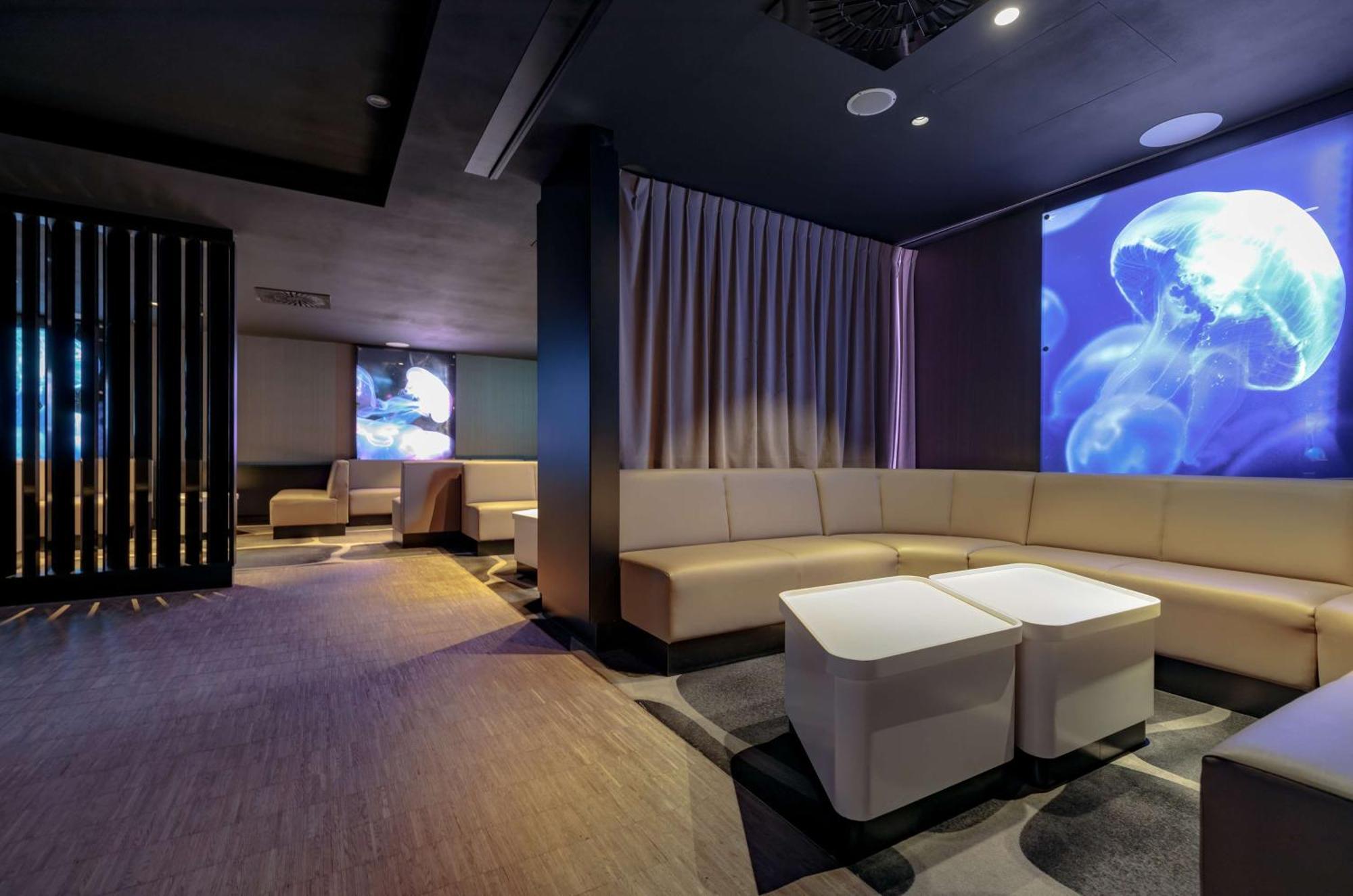 Radisson Blu Resort 슈비노우이스쳬 외부 사진 The photo shows a modern interior space that appears to be a lounge or relaxation area. The design is sleek and minimalist, featuring a curved, light-colored seating arrangement with low tables at the center. The walls are dark, contributing to a coz