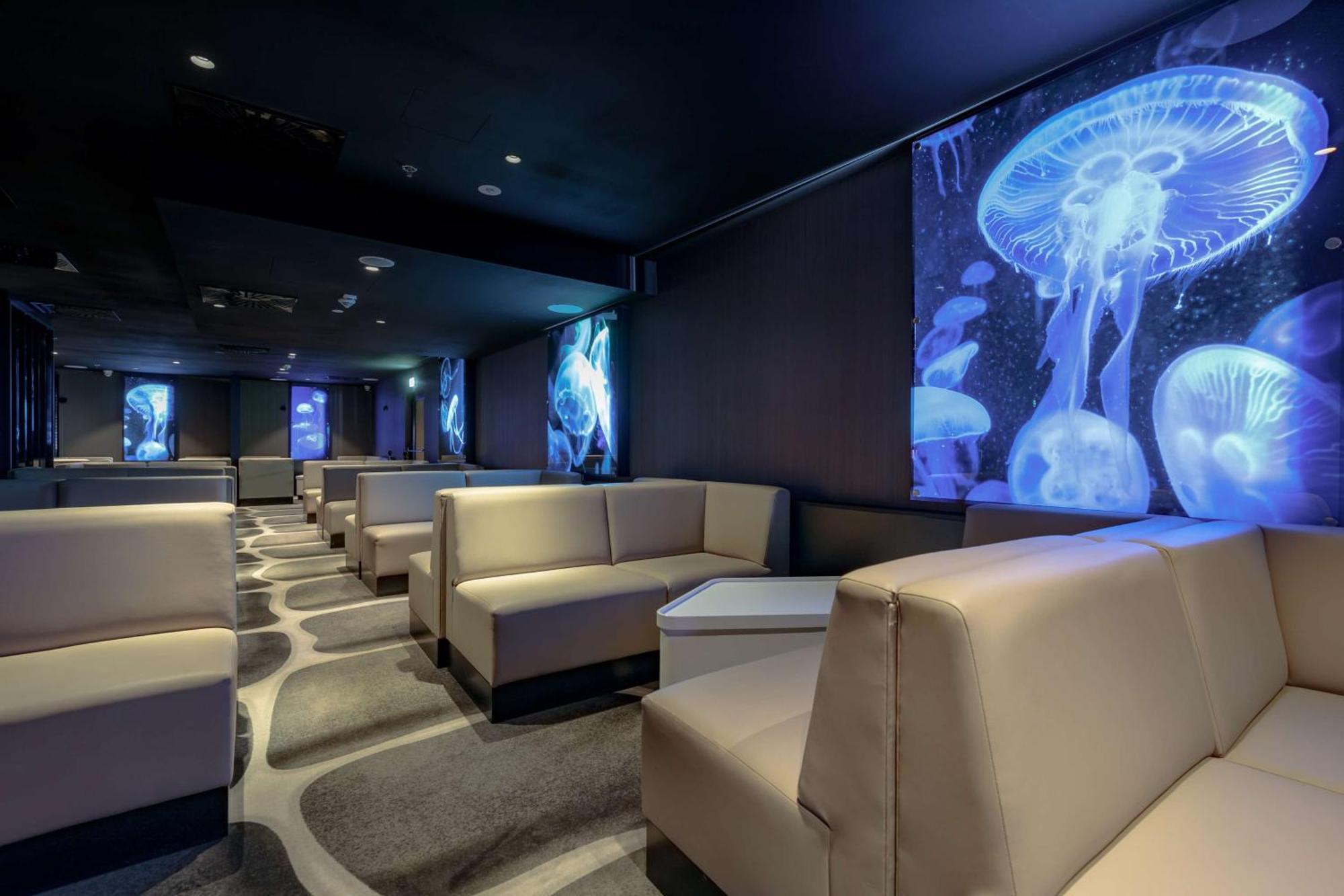 Radisson Blu Resort 슈비노우이스쳬 외부 사진 The photo shows a modern lounge space with a sleek design. The area features light-colored, upholstered seating arranged in an open layout. The walls are decorated with large, vibrant images of jellyfish illuminated in blue, creating a calming underw
