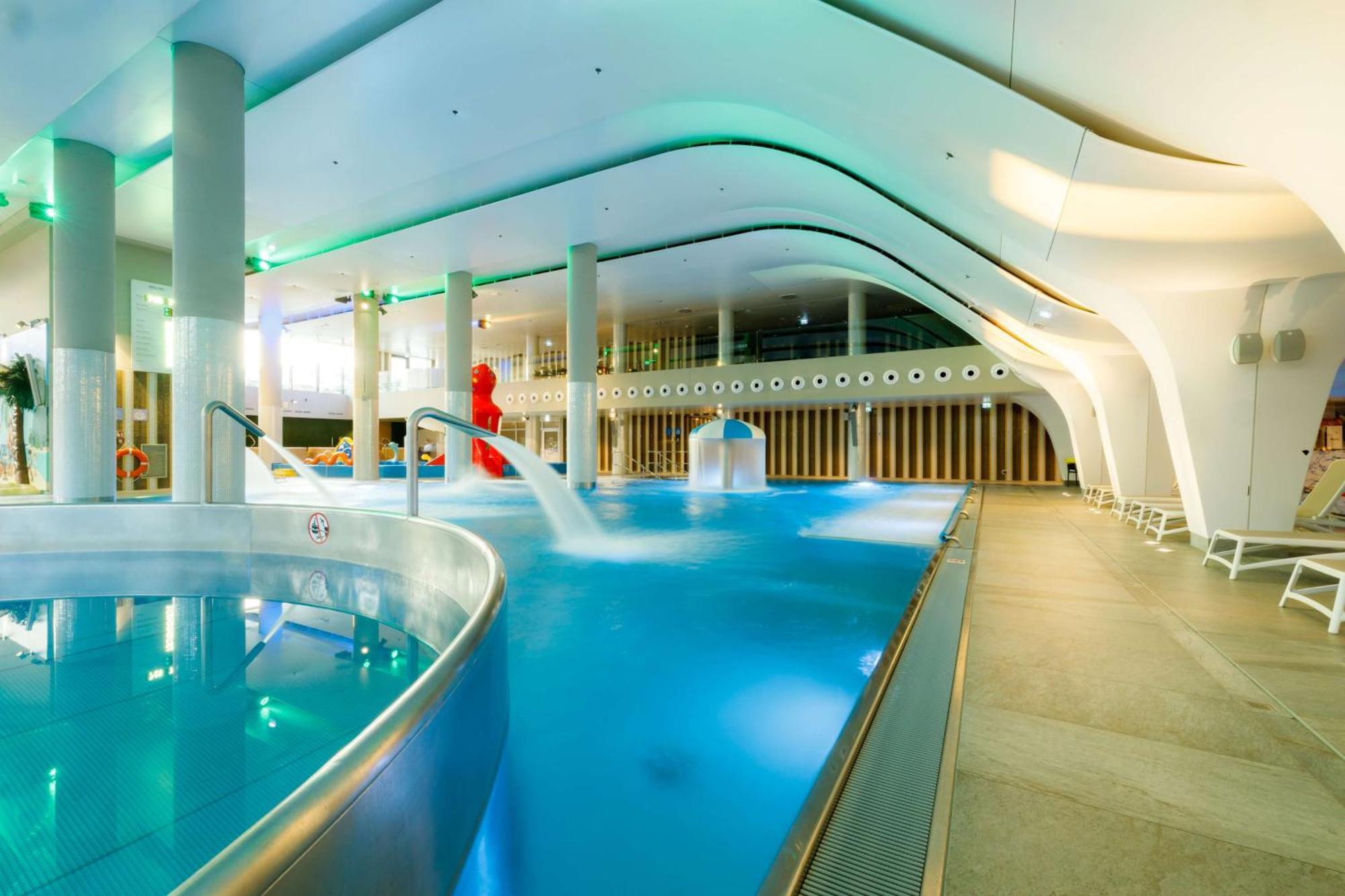 Radisson Blu Resort 슈비노우이스쳬 외부 사진 The photo shows a modern indoor swimming pool area with an elegant design. The pool features curved edges and is filled with clear, blue water. There are various amenities, such as jets for relaxation and possibly seating areas along the edges. The l