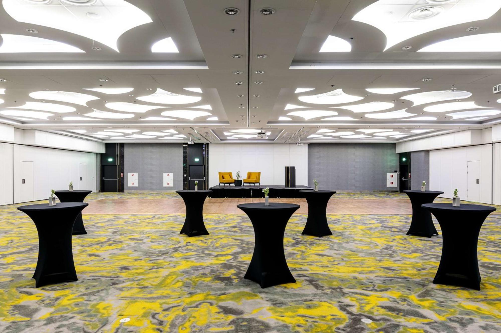 Radisson Blu Resort 슈비노우이스쳬 외부 사진 The photo shows a spacious interior room set up for an event. The flooring features a patterned design in gray with yellow accents. There are several black tall tables scattered throughout the area, and in the background, there are two large yellow a