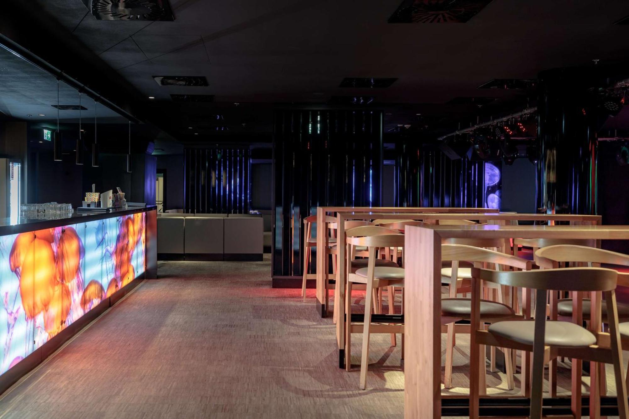 Radisson Blu Resort 슈비노우이스쳬 외부 사진 The photo shows the interior of a modern bar or nightclub. It features a sleek and stylish design with a long bar counter illuminated by colorful lights. The floor has a neutral carpet, and there are wooden high-top tables with accompanying chairs ar