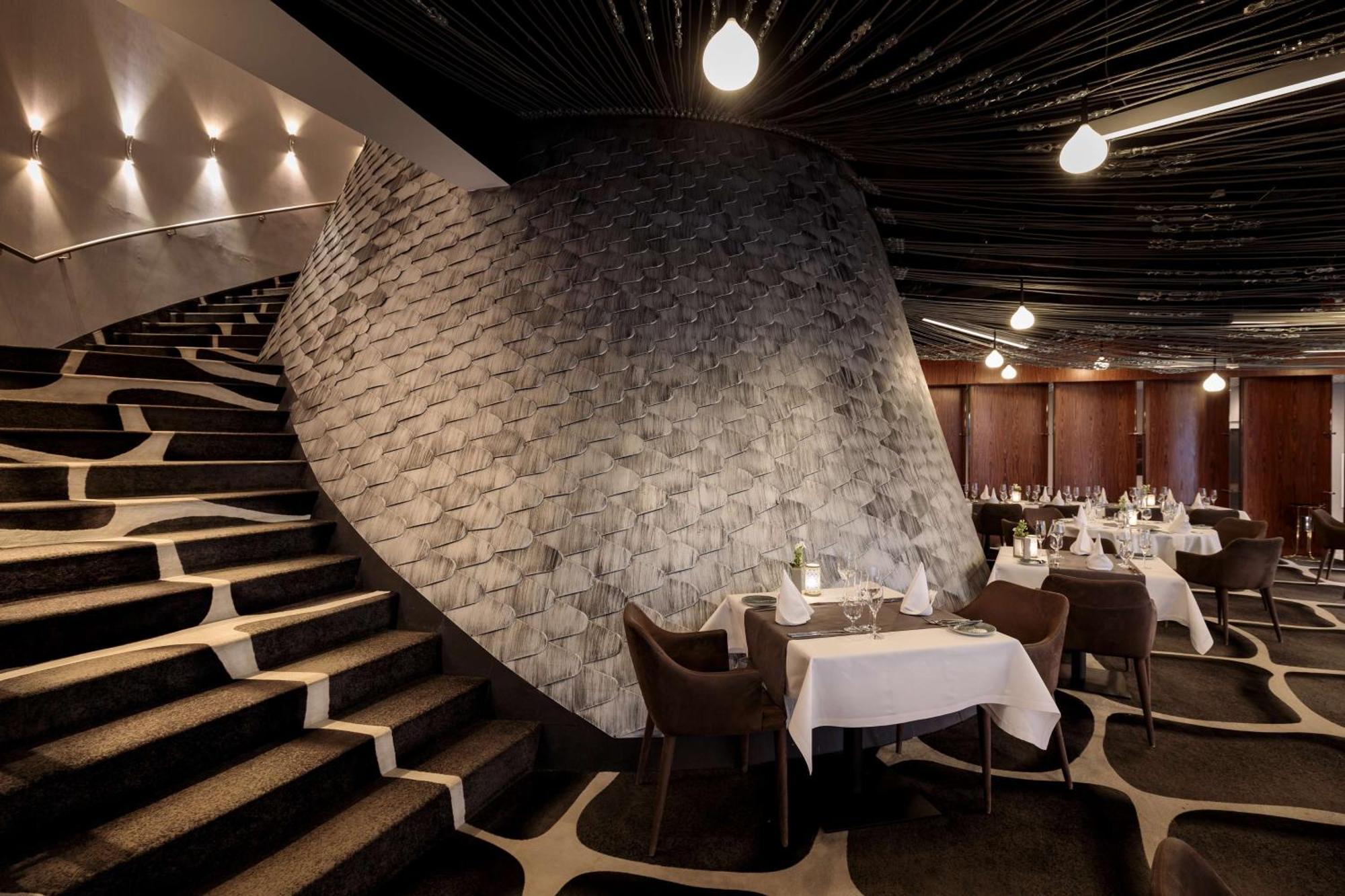 Radisson Blu Resort 슈비노우이스쳬 외부 사진 The photo shows the interior of a stylish restaurant. In the foreground, there are elegantly set tables with white tablecloths and dark chairs. A unique, curving stone wall adds a distinct architectural feature, and a grand staircase with a textured 