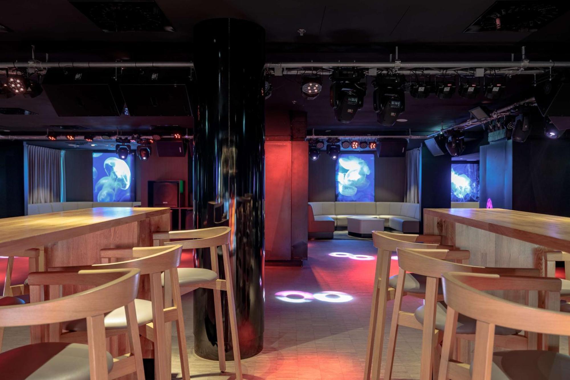 Radisson Blu Resort 슈비노우이스쳬 외부 사진 The photo shows an interior of a modern venue, likely a bar or club. The space features a central black column and light-colored wooden bar stools arranged around a bar area. On the walls, there are large screens displaying vibrant visuals, possibly 