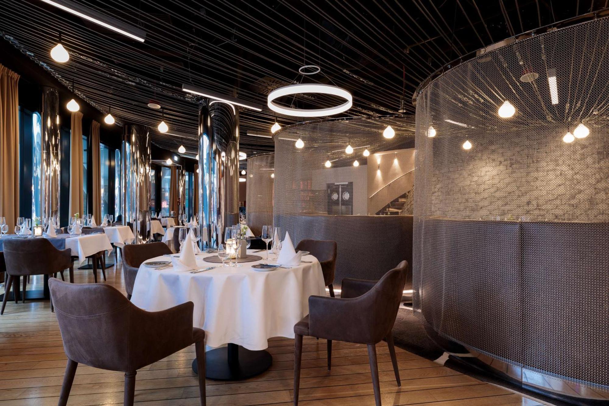 Radisson Blu Resort 슈비노우이스쳬 외부 사진 The image shows a modern restaurant interior that features a stylish design. There are several round tables with white tablecloths. The seating consists of brown, upholstered chairs. The space is illuminated by various hanging lights, including a dis