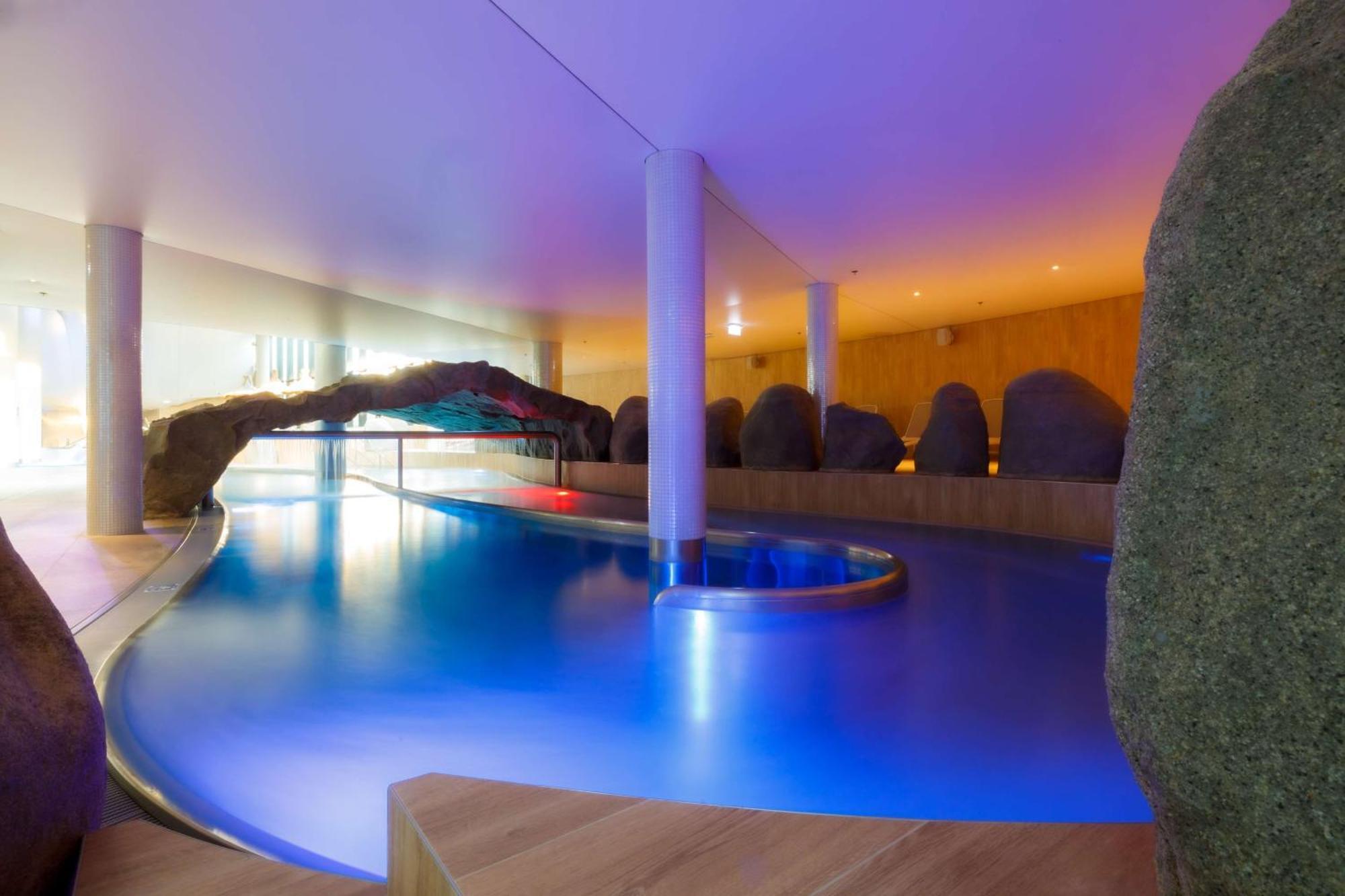 Radisson Blu Resort 슈비노우이스쳬 외부 사진 The photo showcases an elegant indoor swimming pool area. The pool has a flowing, organic shape and is illuminated with soft blue and purple lighting, creating a calming ambiance. Surrounding the pool are smooth, natural-looking stones and wooden ele